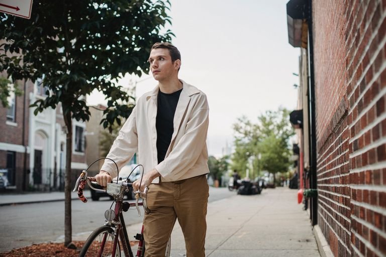 Confident young male in casual outfit strolling on sidewalk with bicycle near road buildings in city street in daylight under bright sky