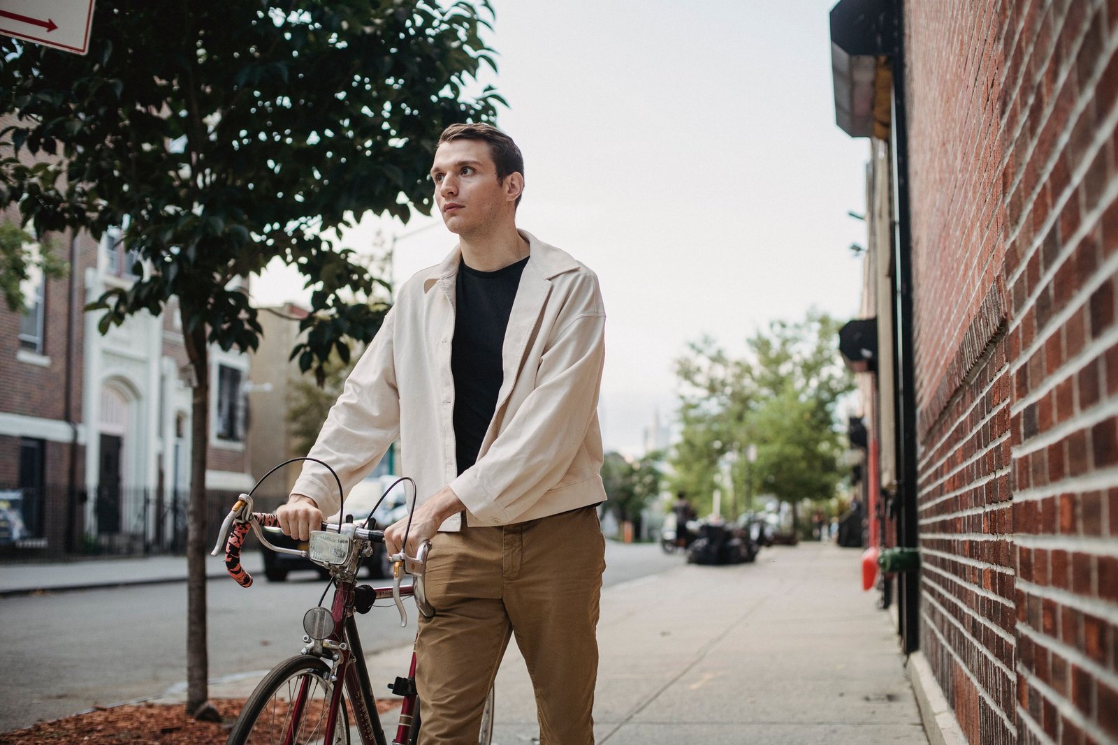 Confident young male in casual outfit strolling on sidewalk with bicycle near road buildings in city street in daylight under bright sky